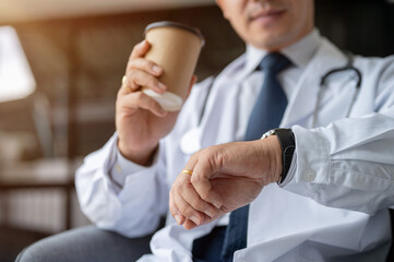 A senior male doctor is checking time on his wristwatch while taking a coffee break in his office.