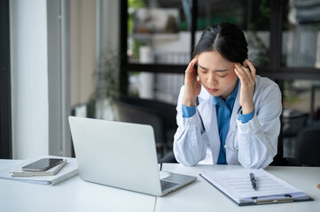 A tired and overworked Asian female doctor is suffering from headache while working at her desk.