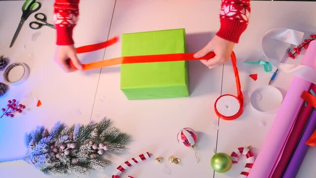 New year DIY gift packing, timelapse, woman hands in red sweater wrap Christmas gift box in green paper, on white table with decor elements, berries, cones, scissors, fir branches, close up. Top view.