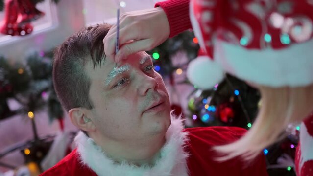 Close-up make-up of young man in image of Santa Claus, girl applying glue on the eyebrows, side view, preparing for Christmas holidays and New Year against backdrop of festive Xmas decoration.