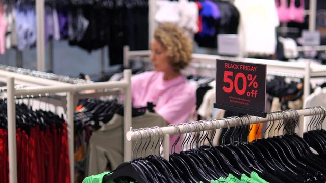 Black Friday signboard in clothing store and customer choosing clothes on background. Wide shot of clothes with sale 50 percent off, stand in store Black Friday. Black Friday shopping evening sales