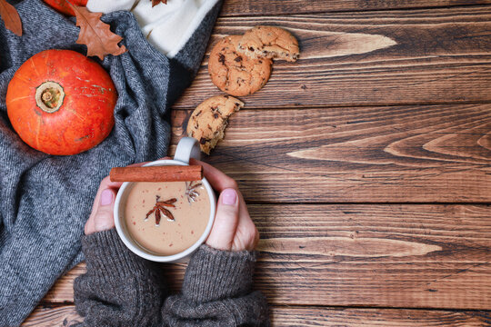 Pretty Female Hands Warming Fresh Coffee Mug, Latte, Spicy Cortado, Bright Orange Pumpkin Wrapped In Woolen Scarf, Sweet Biscuits, Dry Oak Leaves, Spices On Rustic Wooden Table. Autumn Background