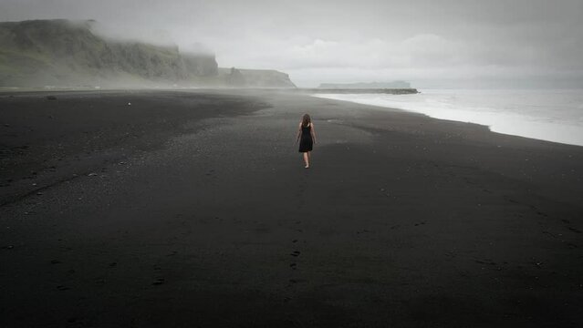 Young beautiful woman in black dress walking on black sand beach Iceland, foggy dramatic mountain landscape, tracking shot