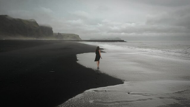 Young beautiful woman in black dress running on black sand beach Iceland through waves, aerial tracking shot