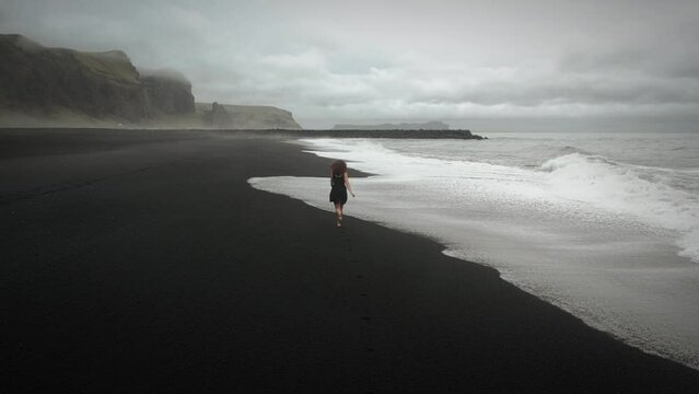 Aerial young beautiful woman in black dress running on black sand beach Iceland, tracking shot	