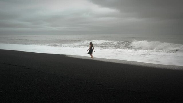 Young beautiful woman in black dress running on black sand beach Iceland, aerial dramatic waves seascape