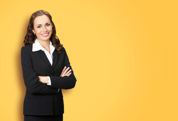Portrait image of happy smiling young business woman standing in crossed arms pose, black confident suit, isolated yellow color background. Female executive office worker, teacher or real estate agent