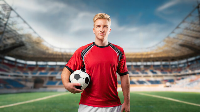 Caucasian young blonde male soccer player holding a soccer ball with stadium background.
