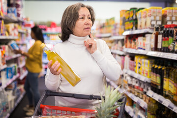 Portrait of pensive elderly woman making purchases in supermarket, choosing vegetable oil