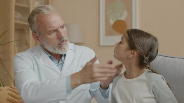 Medium shot of professional Caucasian male doctor touching neck of Asian little girl while examining thyroid glands, doing checkup appointment at homeMedium shot of professional Caucasian male doctor 