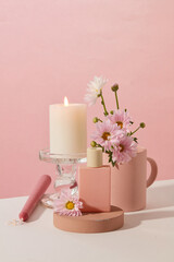 Obraz na płótnie Canvas A burning candle is placed on a glass candlestick, an unlabeled perfume bottle is placed on a wooden podium and fresh flowers are on a pink and white background. Copy space for ads.