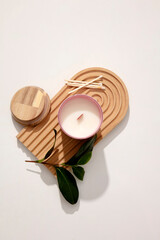 A jar of scented candles is placed on a domed wooden platform with cotton swabs and green leaves on a white background. Enjoy relaxing moments with the scent of candles.