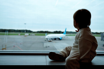 Interested toddler girl sits on windowsill of panoramic window in airport terminal. Silhouette of...