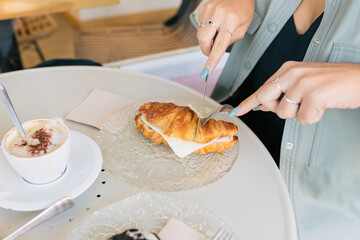 Crop woman eating croissant and taking coffee in cafe