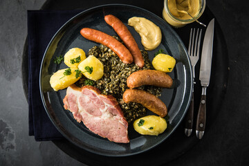 Oldenburger Grünkohl mit geräucherter Kohlwurst, Pinkel, Kassler Nacken, Kartoffeln, Senf, Petersilie auf Teller und dunkel schwarz Schiefer Hintergrund