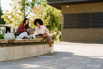 Two female students discussing something while studying together sitting on bench at street