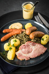 Oldenburger Grünkohl mit geräucherter Kohlwurst, Pinkel, Kassler Nacken, Kartoffeln, Senf, Petersilie auf Teller und dunkel schwarz Schiefer Hintergrund