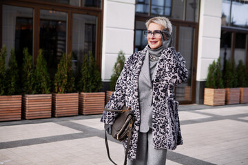 Stylish cool mature woman model wears fashionable winter multilayer outfit, animal print coat, grey wool suit with turtleneck and eyeglasses, stands in city. Elder Female in Trendy clothes