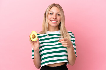 Young caucasian woman holding an avocado isolated on pink background surprised and pointing front