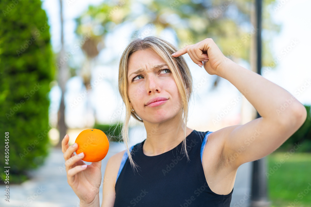 Wall mural young blonde sport woman holding an orange at outdoors having doubts and with confuse face expressio