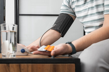 Obraz premium Unrecognizable man sitting on a sofa at home and measures blood pressure on a hand with modern digital tonometer and holding blister packs with pills, medicines