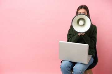 Young caucasian woman sitting on a chair with her laptop isolated on pink background shouting...