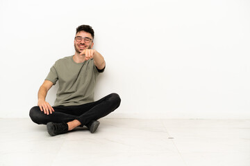 Young caucasian man sitting on the floor isolated on white background points finger at you with a confident expression