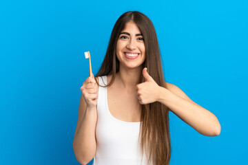 Young caucasian woman brushing teeth isolated on blue background giving a thumbs up gesture