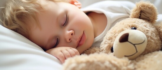 Sweet baby boy in bear overall, sleeping in bed with teddy bear - obrazy, fototapety, plakaty