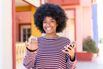 African American girl using mobile phone at outdoors inviting to come with hand. Happy that you came