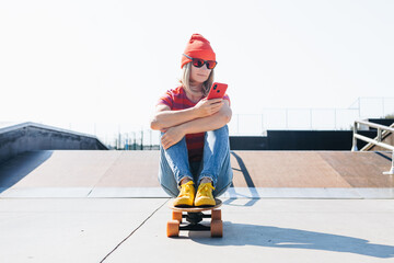 Beautiful young hipster woman using smart phone On Skateboard at summer day. Active lifestyle, modern life, subculture concept