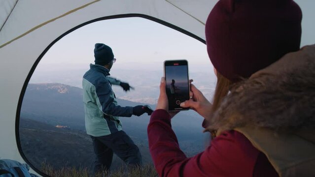 Young woman takes a photo of tourist on beautiful mountain scenery using phone. African American hiker with axe in hands poses on hill. Backpacker friends stopped to rest during adventure vacation.