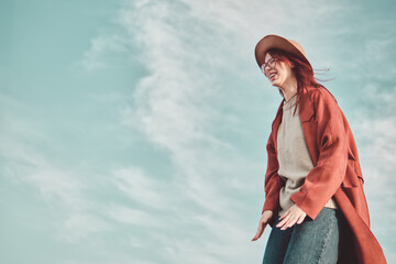 Laughing teenage girl in red coat and hat standing against sky background. Low angle view. © TanyaJoy