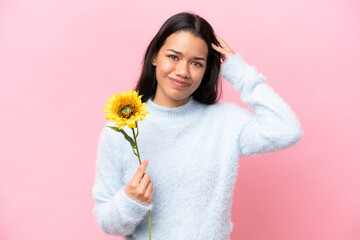 Young Colombian woman holding sunflower isolated on pink background having doubts