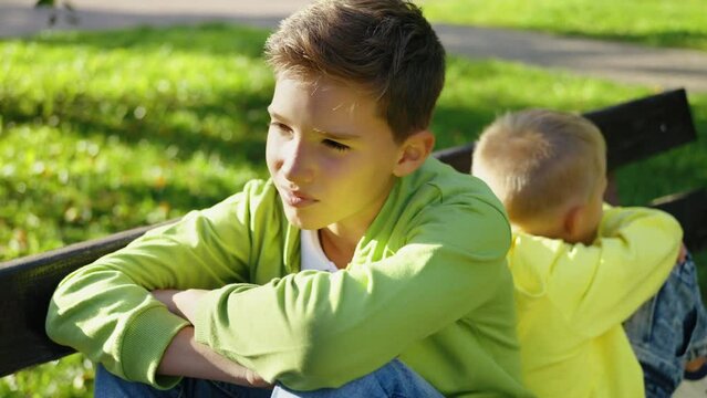 Two brothers sitting back to back on bench in park after argument. Boys are angry at each other. Siblings holding grudge after quarrel, no one wanted to give in.