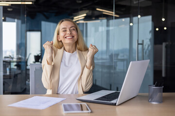 Close-up portrait of a happy business woman who is close eyes happy about success and achievement, sitting at the desk in the office and showing a victory gesture with her hands, happy for the camera