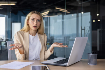 Portrait of an upset and disappointed businesswoman sitting in the office at the table, working on a laptop, looking seriously at the camera and spreading her hands, thinking about solving a problem