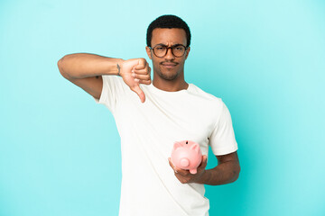 African American handsome man holding a piggybank over isolated blue background showing thumb down...
