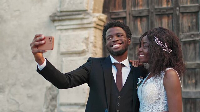 African American couple making photo on phone while standing before old building. Young husband hugging his wife while doing selfie together. Loving people smiling and feeling happy with each other.