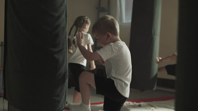 Child attacks punching bag with classmates in gym. Toddler boy beats hanging equipment by legs and hands in classroom slow motion. Kickboxing team