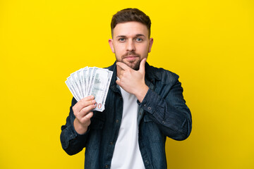 Young caucasian man taking a lot of money  isolated on yellow background thinking