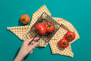 Hand of woman holding a black basket containing fresh tomatoes with checkered cloth on green background. Scene for advertising, natural ingredient for beauty product