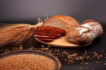 Whole and sliced breads, wheat ears and scattered wheat grains on wooden plate on black background....