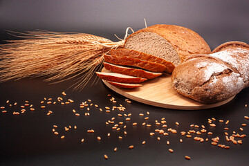 Whole and sliced breads, wheat ears and scattered wheat grains on wooden plate on black background....