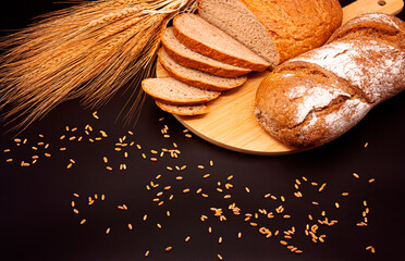 Whole and sliced breads, wheat ears and scattered wheat grains on wooden plate on black background. Photograph.