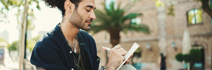 Young italian guy with ponytail and stubble sits on street bench and makes sketches with pen on...