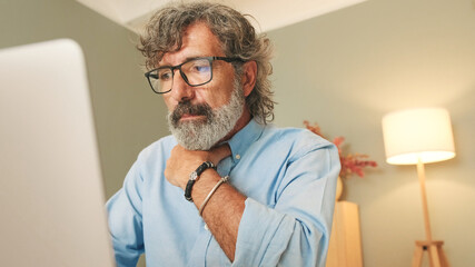 An elderly man with gray hair wearing glasses sits in the living room in modern apartment working...