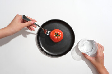 Female hand model holding a fork and a glass of water. A tomato placed on black dish. The Vitamin A...