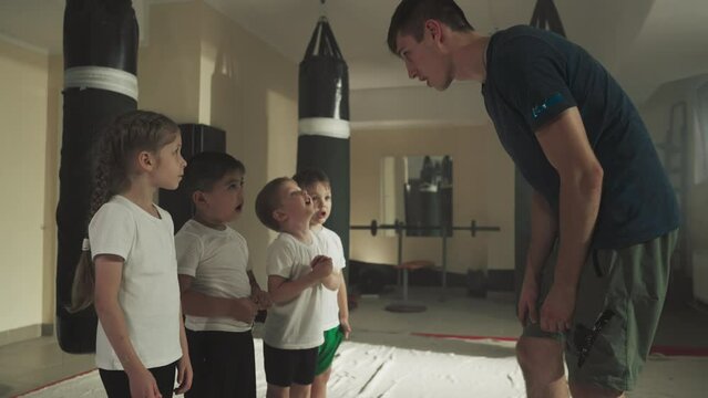 Children look at trainer showing block in gym. Funny junior students learn fighting technique from instructor in sports club slow motion. Kids team at self-defense lesson