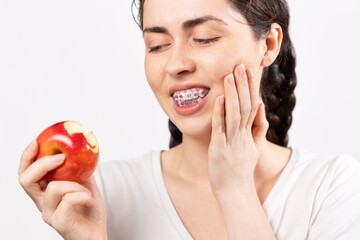 Portrait of young Caucasian woman with braces suffering from pain because bit off red apple. White background. Concept of forbidden food during orthodontic treatment